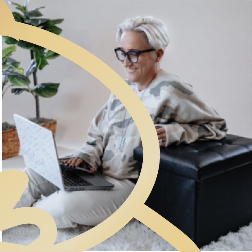 Woman with short gray hair and glasses sitting on a bench, smiling while using a laptop in a bright indoor space with plants nearby.
