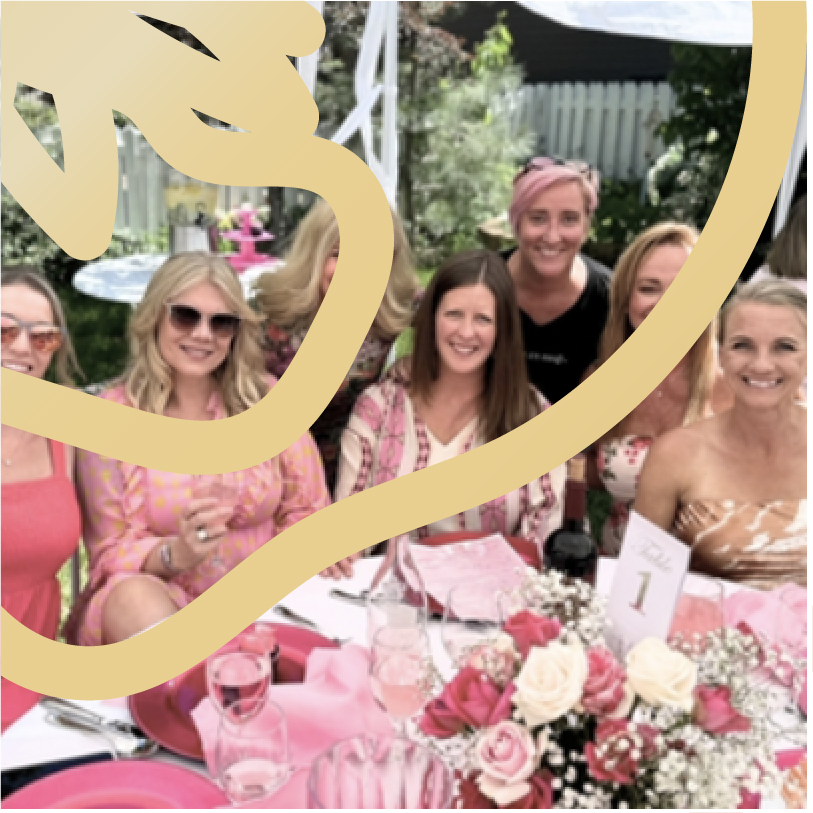 Group of women smiling at a decorated outdoor table with pink flowers and table settings, gathered for a celebration.