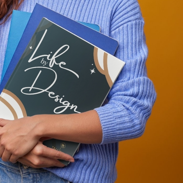 Person wearing a blue sweater holding notebooks, including one titled “Life by Design” with a decorative cover design.
