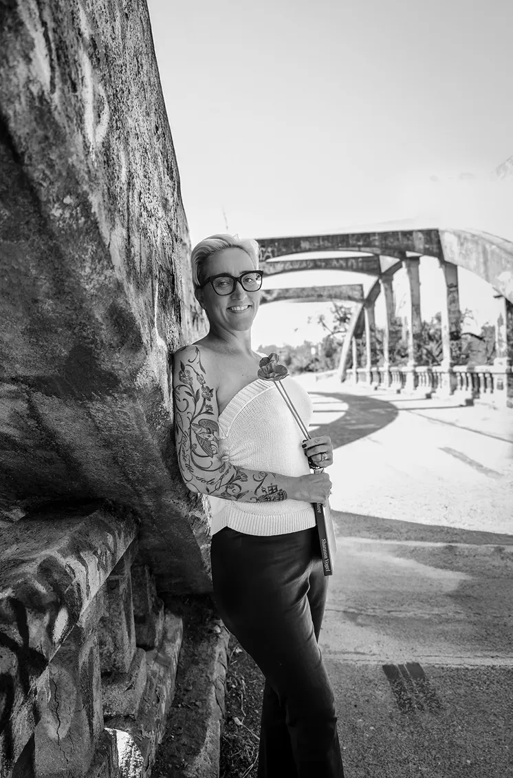Black-and-white photo of a woman with glasses leaning against a stone wall near an arched bridge, smiling at the camera.