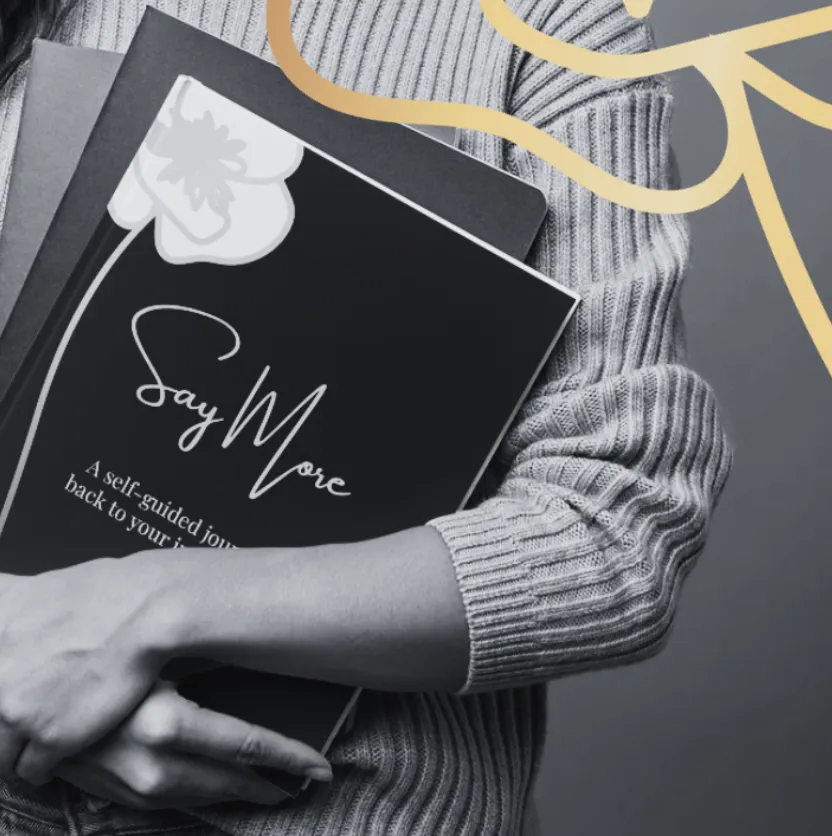 Black-and-white close-up of a person holding a “Say More” workbook with a poppy illustration, partially overlaid with a gold graphic element.