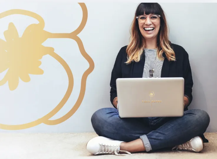 Smiling woman sitting cross-legged on the floor with a laptop featuring the “Tall Poppy” logo, against a wall with a large poppy illustration.