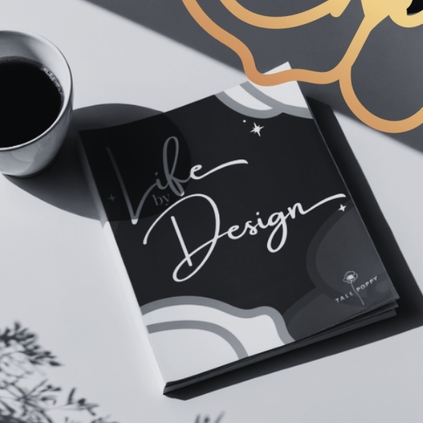 Black-and-white photo of a “Life by Design” workbook on a table beside a cup of coffee, with a gold graphic overlay in the corner.