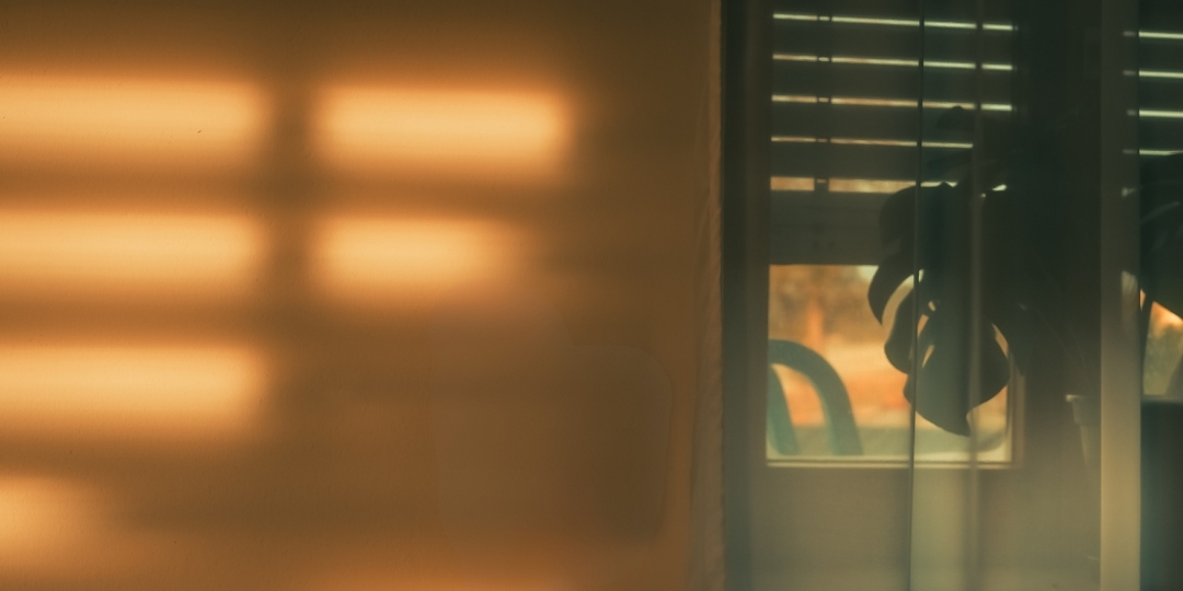 Soft golden light streaming through window blinds, casting warm shadows on a wall with a faint silhouette visible in the background.