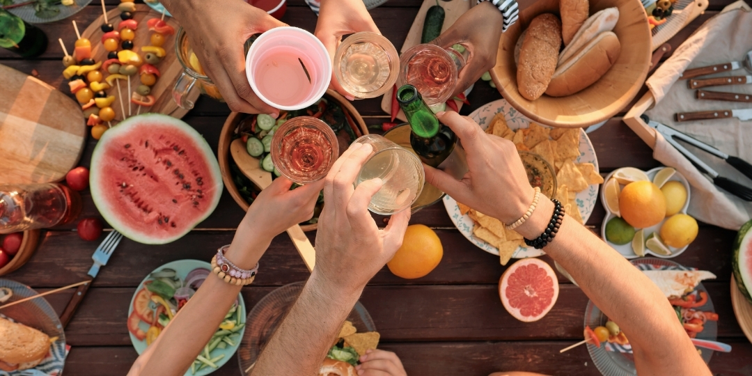 Overhead view of friends clinking glasses in a toast over a wooden table filled with fresh fruit, bread, and summer foods.