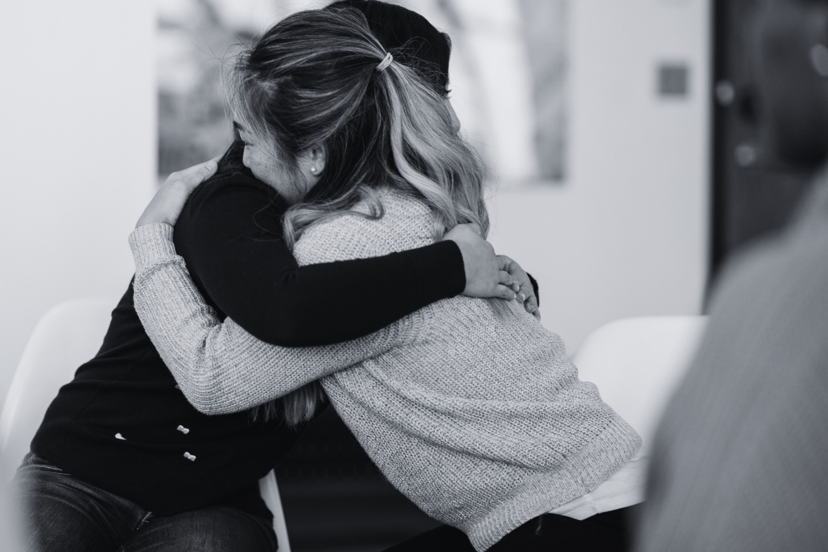 Black-and-white photo of two women sitting and sharing a supportive hug indoors.