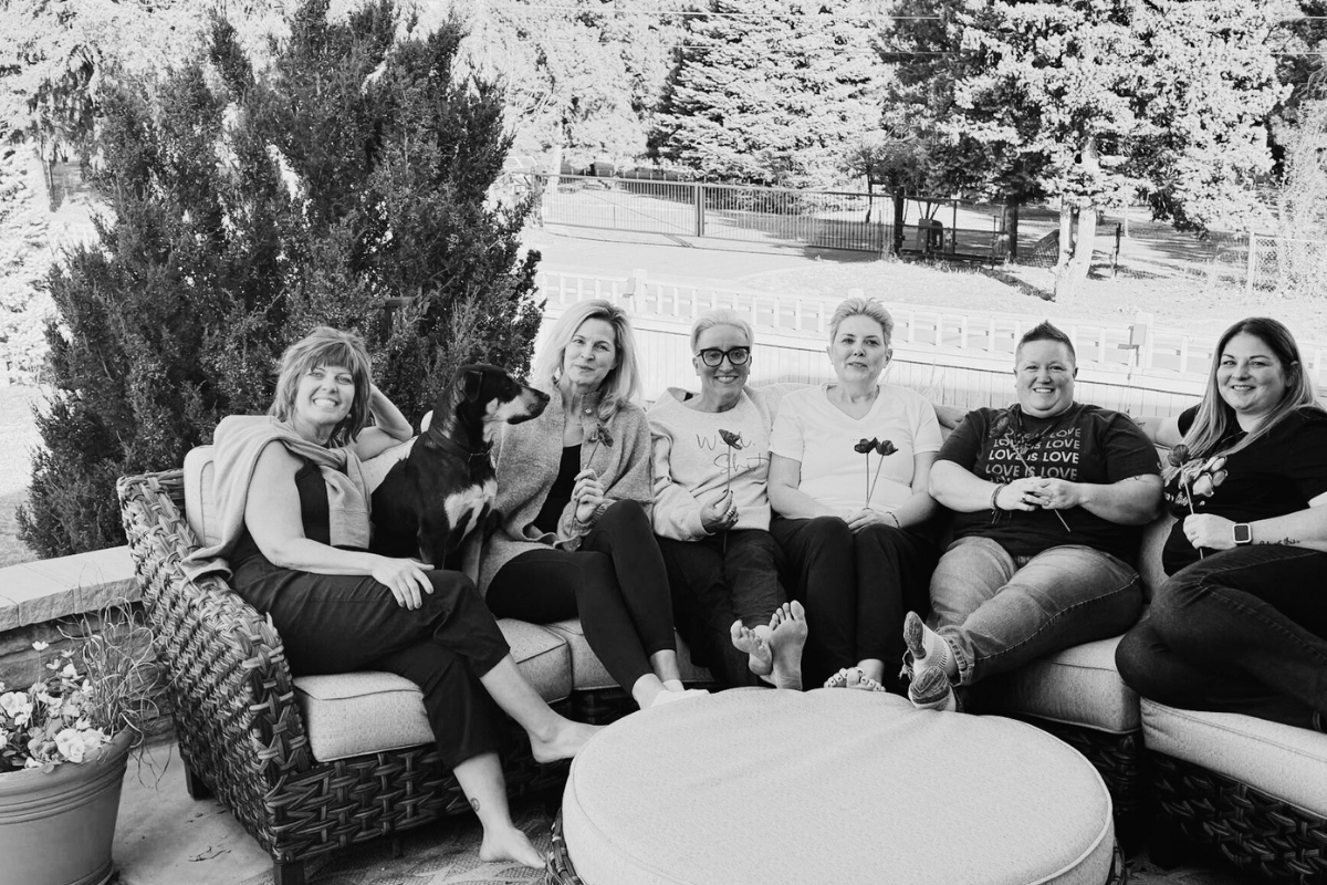 Black-and-white photo of five women sitting together on an outdoor patio couch, smiling and relaxing.