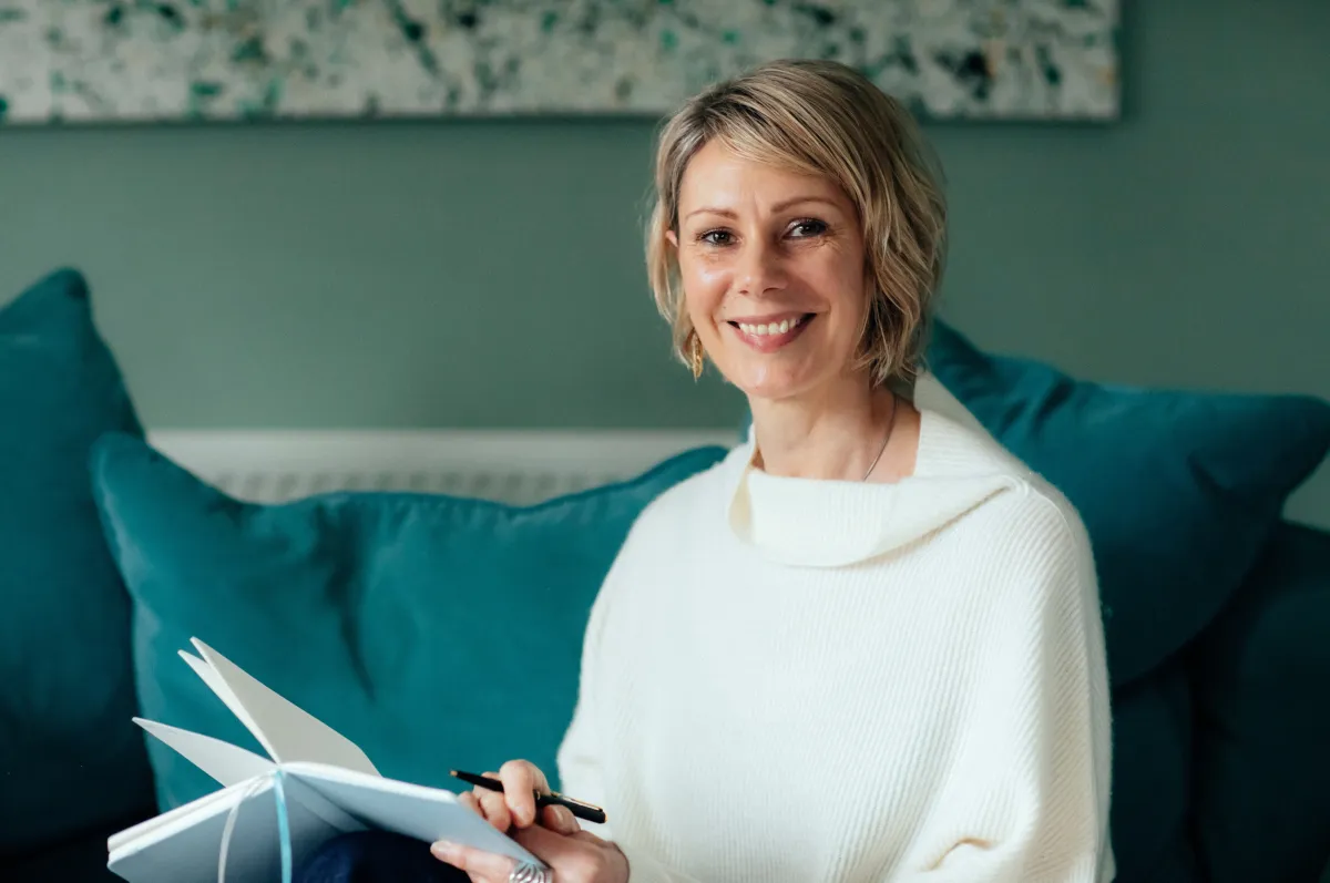 Kate sitting on a teal sofa with a white jumper, smiling at the camera, holing an open notebook