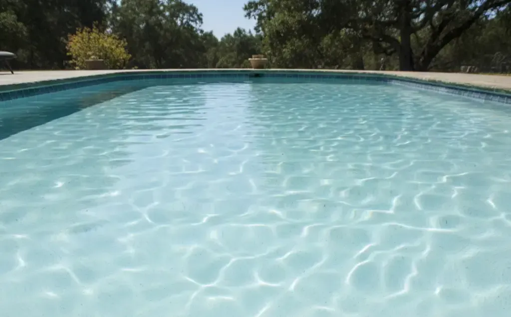 Pool cleaning tools and water test strips on a table beside a cloudy backyard swimming pool, showing pool maintenance and water chemistry testing