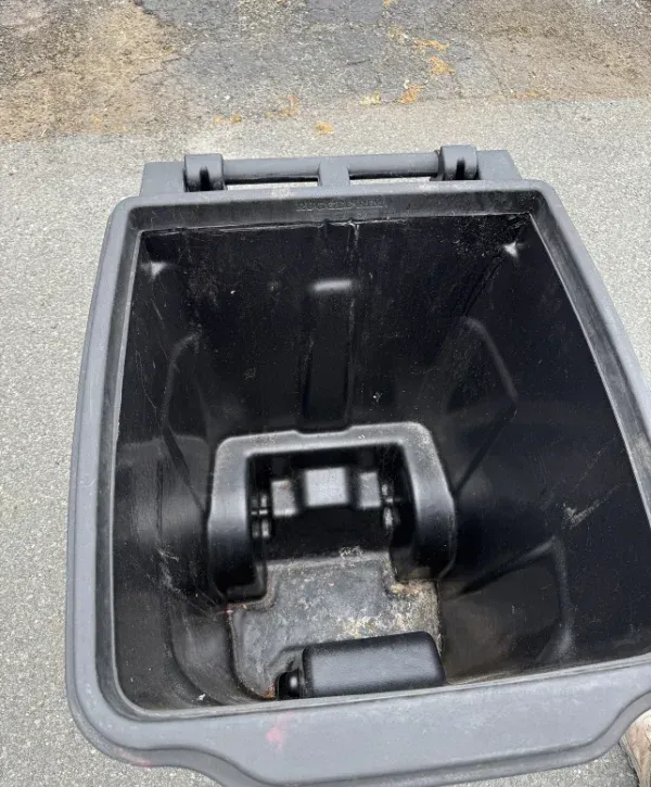An overhead view looking into an empty, dirty black plastic garbage can outdoors on asphalt.