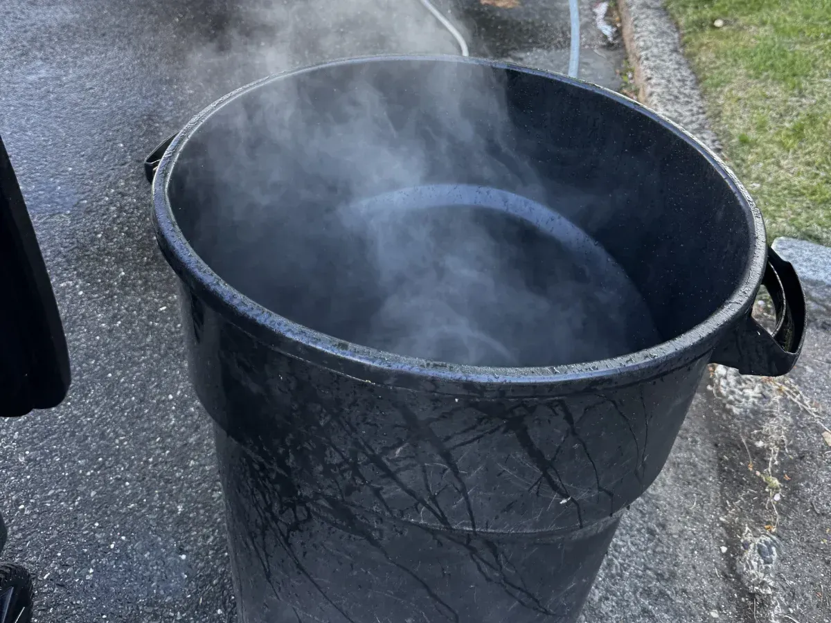 A black plastic barrel filled with hot water from which steam is rising, sitting on asphalt.