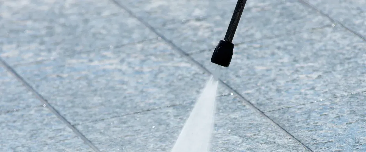 A close-up of a pressure washer nozzle spraying water onto light blue concrete patio tiles.