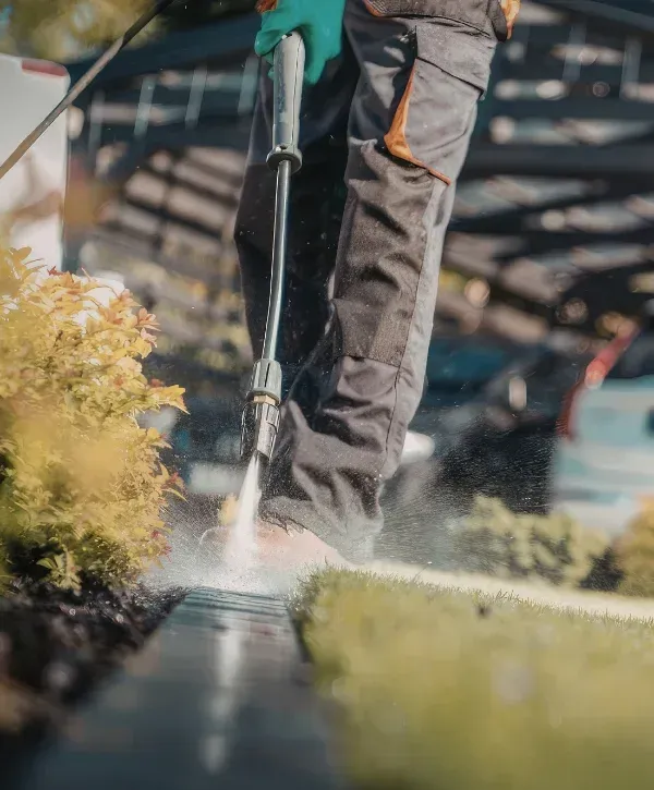 A person in work pants and green gloves pressure washing a black drainage grate alongside a lawn.