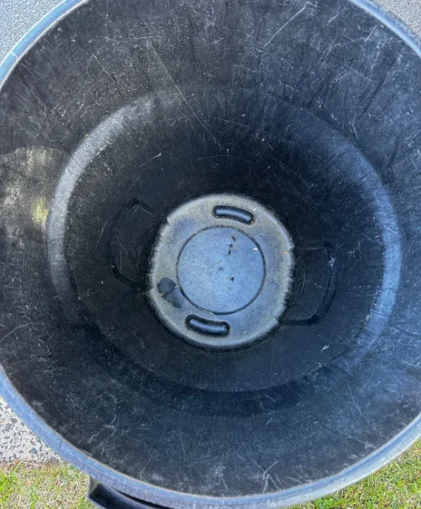Looking straight down into a dusty, empty black plastic trashcan with scuff marks and some liquid at the bottom.