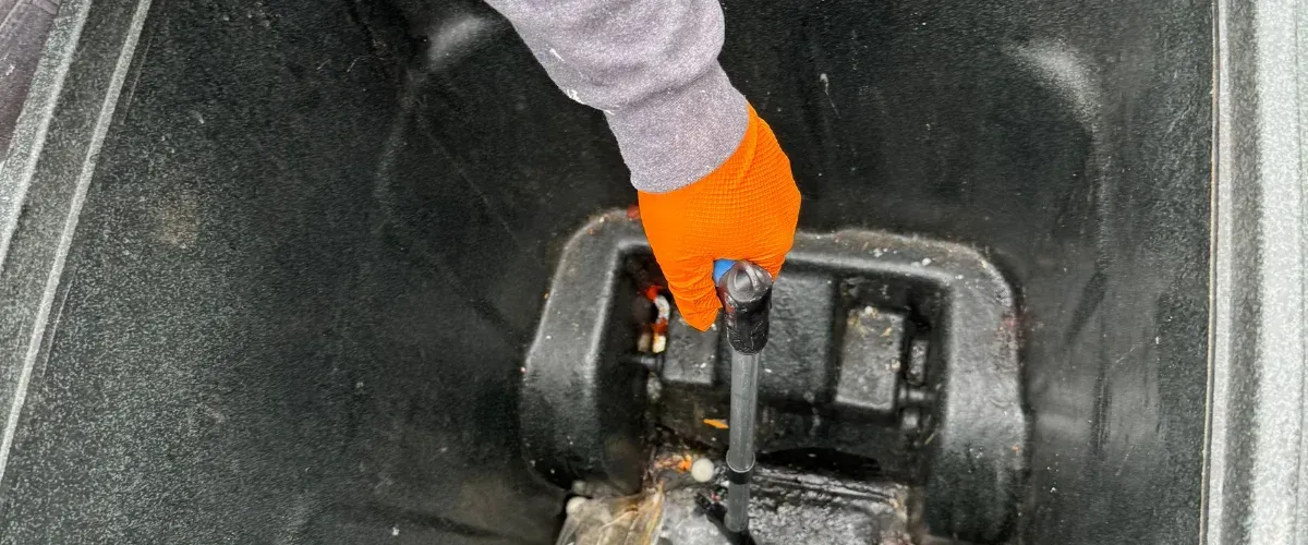 A person's hand in an orange glove holds a black pump handle in a dark, dirty well or container.