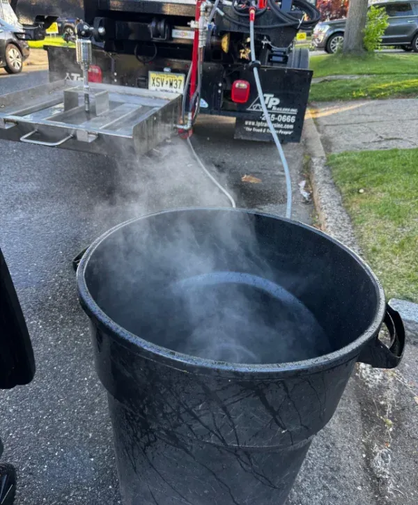 A black industrial barrel on asphalt steams with hot water, next to the rear of a black utility truck.