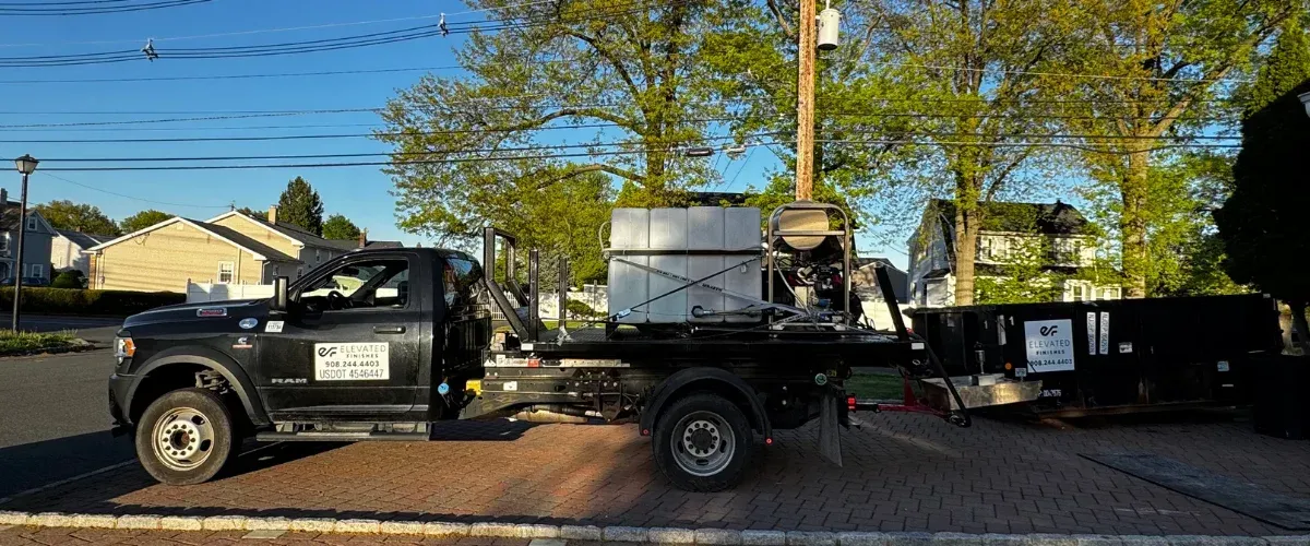 A black Elevated Finishes flatbed truck with a large tank and equipment parked on a brick sidewalk next to a street.