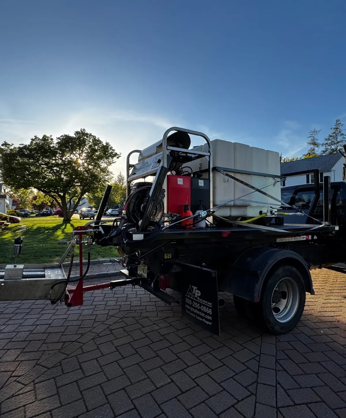 Rear side of a utility truck with equipment, including a white tank and red canister, parked on pavers at sunset.