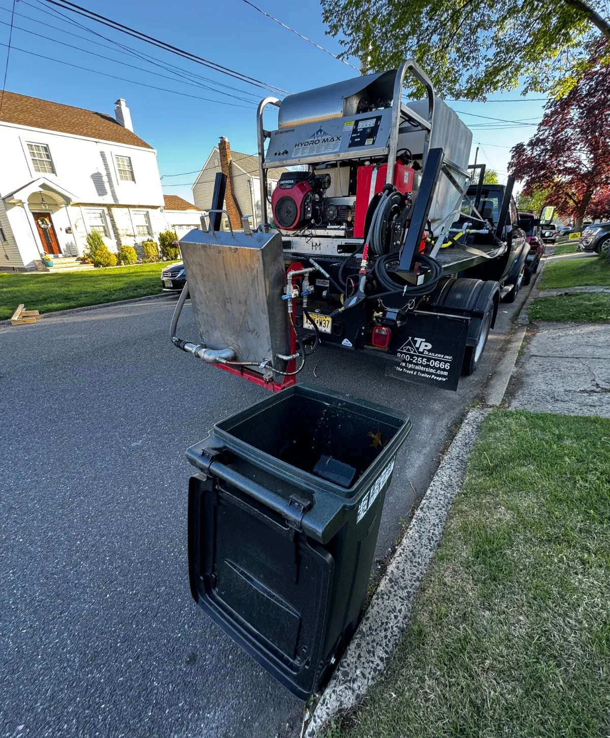 Rear view of a Hydro Max utility trailer with a Honda engine parked on a residential street next to a green trash can.
