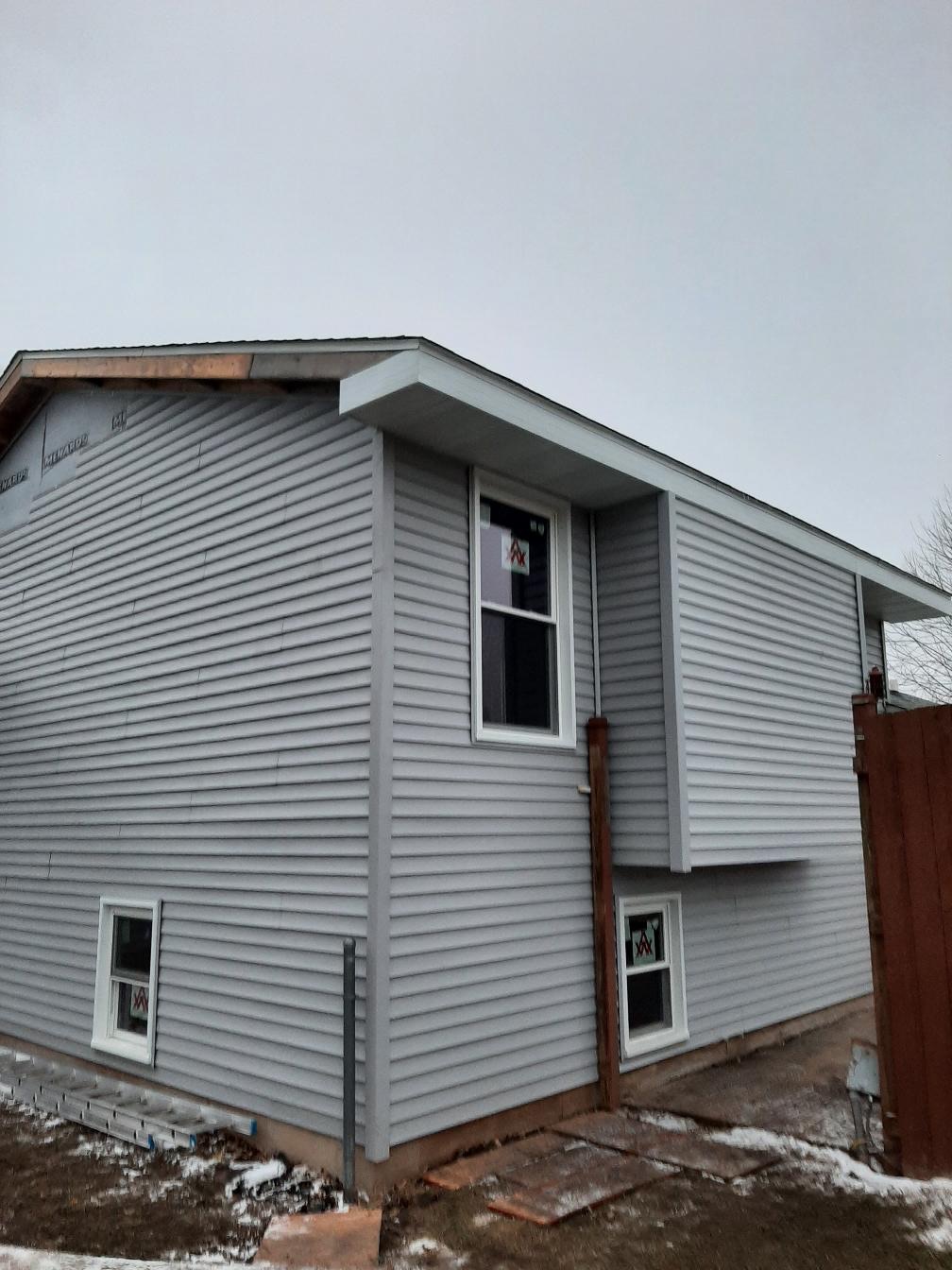 A residential home exterior under construction showcasing newly installed light gray horizontal vinyl siding and white-trimmed windows. Siding installation completed by Precision Exterior Inc., serving Lena, IL and surrounding areas, featuring precise corner vinyl wrapping and soffit integration.