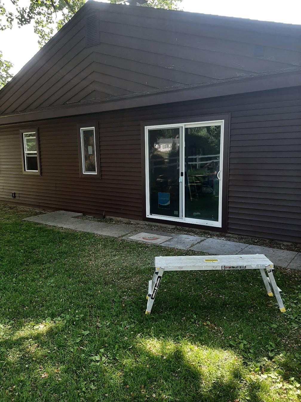 A residential home exterior featuring newly installed dark brown horizontal siding with matching brown trim. The project highlights a white-trimmed sliding glass patio door and two matching single-hung windows seamlessly integrated into the siding by Precision Exterior Inc., serving Lena, IL and surrounding areas.