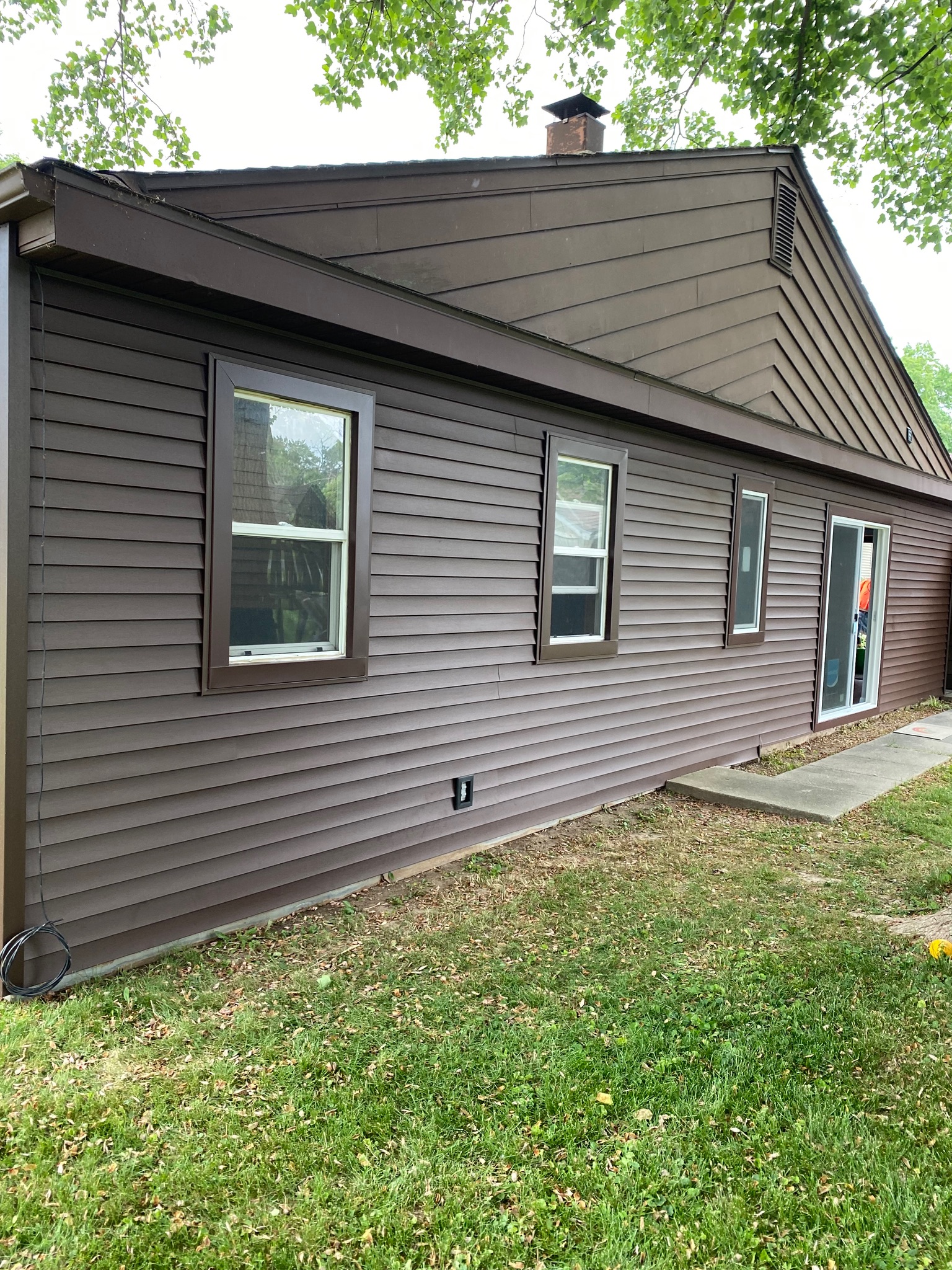 A residential home exterior showcasing completed dark brown horizontal siding with matching trim around the windows and roofline. The project highlights newly installed single-hung windows and a sliding glass patio door, expertly completed by Precision Exterior Inc., serving Lena, IL and surrounding areas.