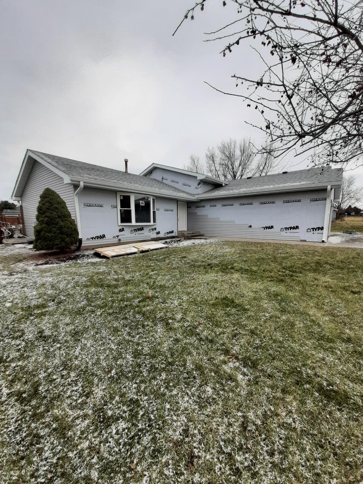 Wide shot of a residential home siding replacement project in progress by Precision Exterior Inc. in Lena, IL. The image shows a completed light gray vinyl siding gable wall on the left, while the main front exterior walls are wrapped in TYPAR weather barrier, showcasing rows of light gray horizontal siding partially installed along the bottom sections under an overcast sky.