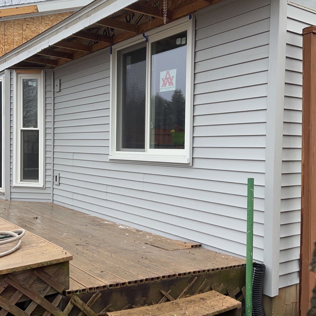 A residential home exterior featuring newly installed light gray horizontal vinyl siding, crisp white trim, and a newly installed white vinyl sliding window overlooking an outdoor wooden deck. Siding and window installation completed by Precision Exterior Inc., serving Lena, IL and surrounding areas.