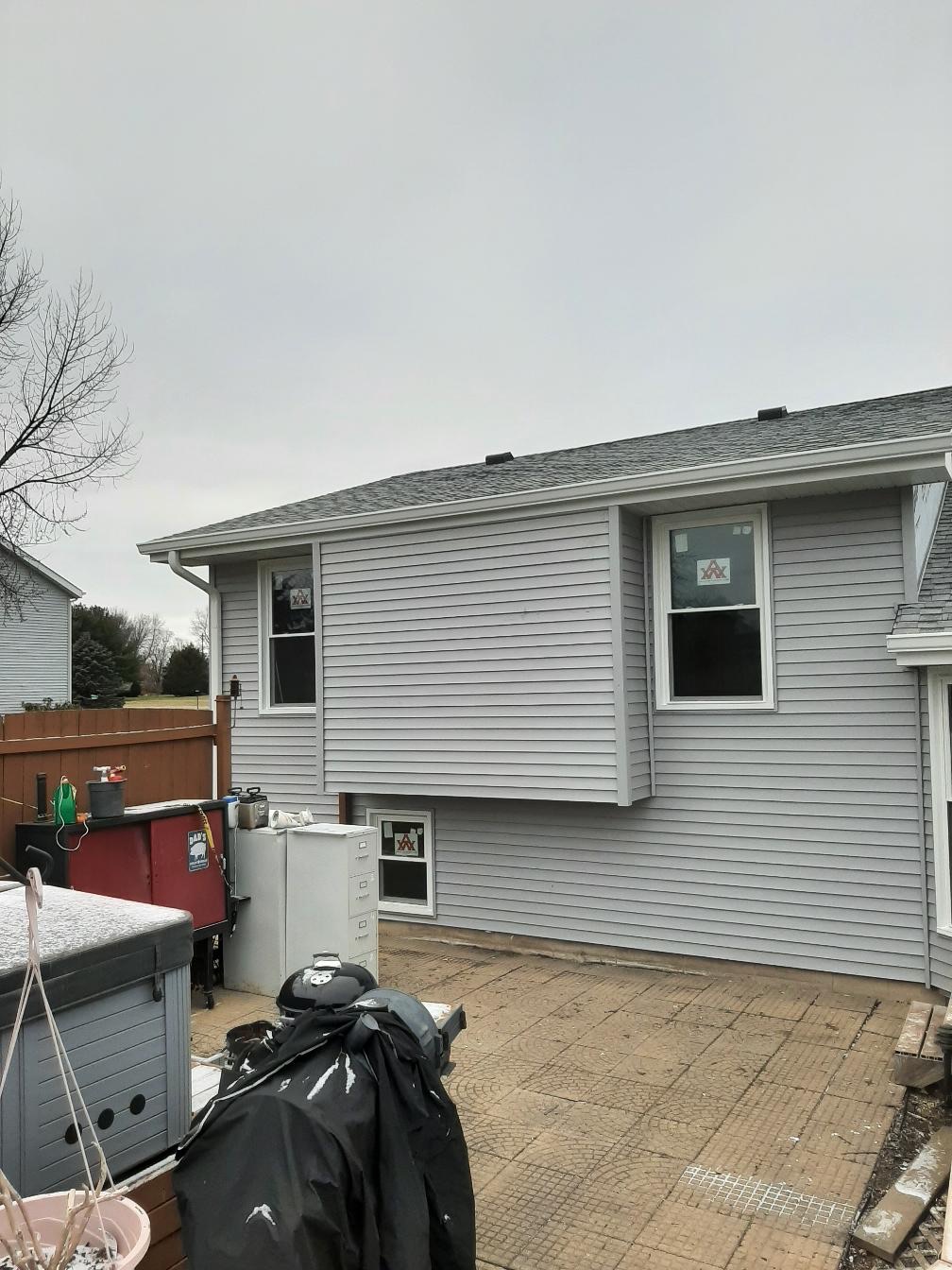 Completed light gray vinyl siding installation on a two-story home exterior by Precision Exterior Inc. in Lena, IL. The image showcases clean siding lines around newly installed double-hung windows, a basement window, and white trim work overlooking a patio area under an overcast sky.