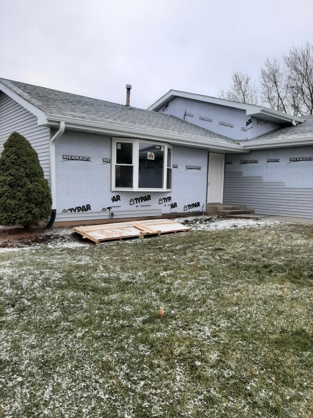 A residential home exterior undergoing a siding replacement project by Precision Exterior Inc. in Lena, IL. The image captures the installation in progress, showing a newly completed light gray vinyl siding gable section alongside front walls covered in TYPAR weather barrier, a new white vinyl bay window, and partial siding installation by the front entryway.