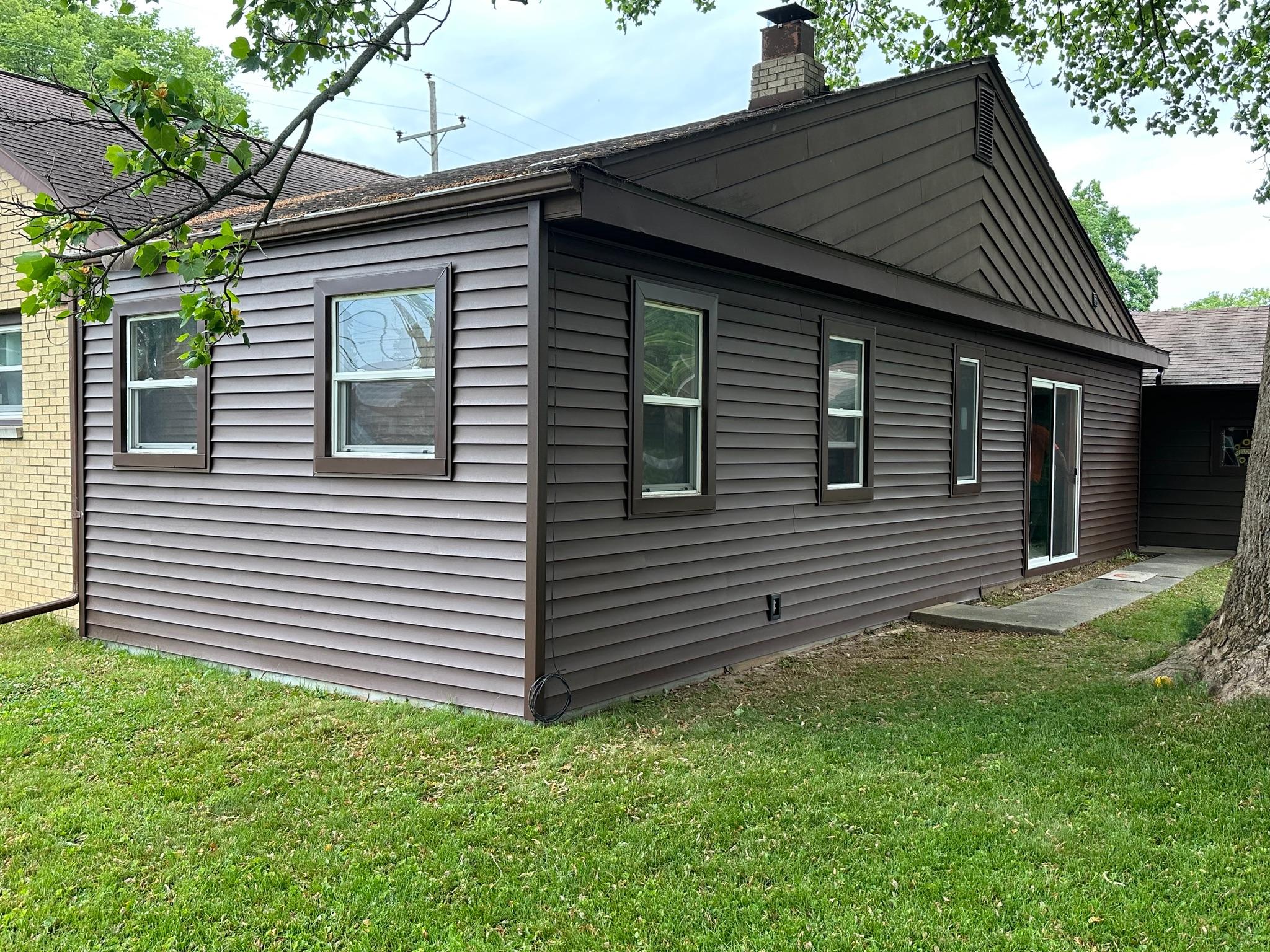 A residential home exterior corner showcasing completed dark brown horizontal vinyl siding with matching dark brown trim around multiple single-hung windows. A white-framed sliding patio door is visible along the side wall, precisely integrated into the updated siding by Precision Exterior Inc., serving Lena, IL and surrounding areas.