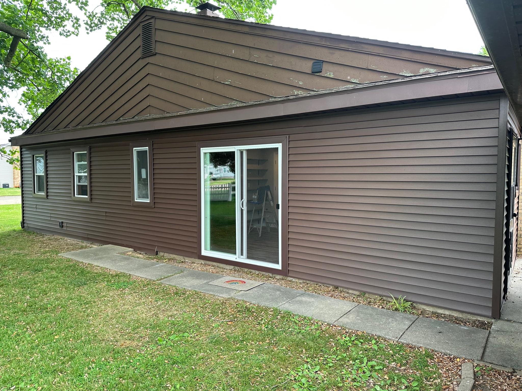 A residential home exterior featuring newly installed dark brown horizontal vinyl siding with matching dark brown trim on the main wall and a contrasting older gable. A white-trimmed sliding glass patio door and multiple matching single-hung windows are seamlessly integrated into the updated siding. Work completed by Precision Exterior Inc., serving Lena, IL and surrounding areas.