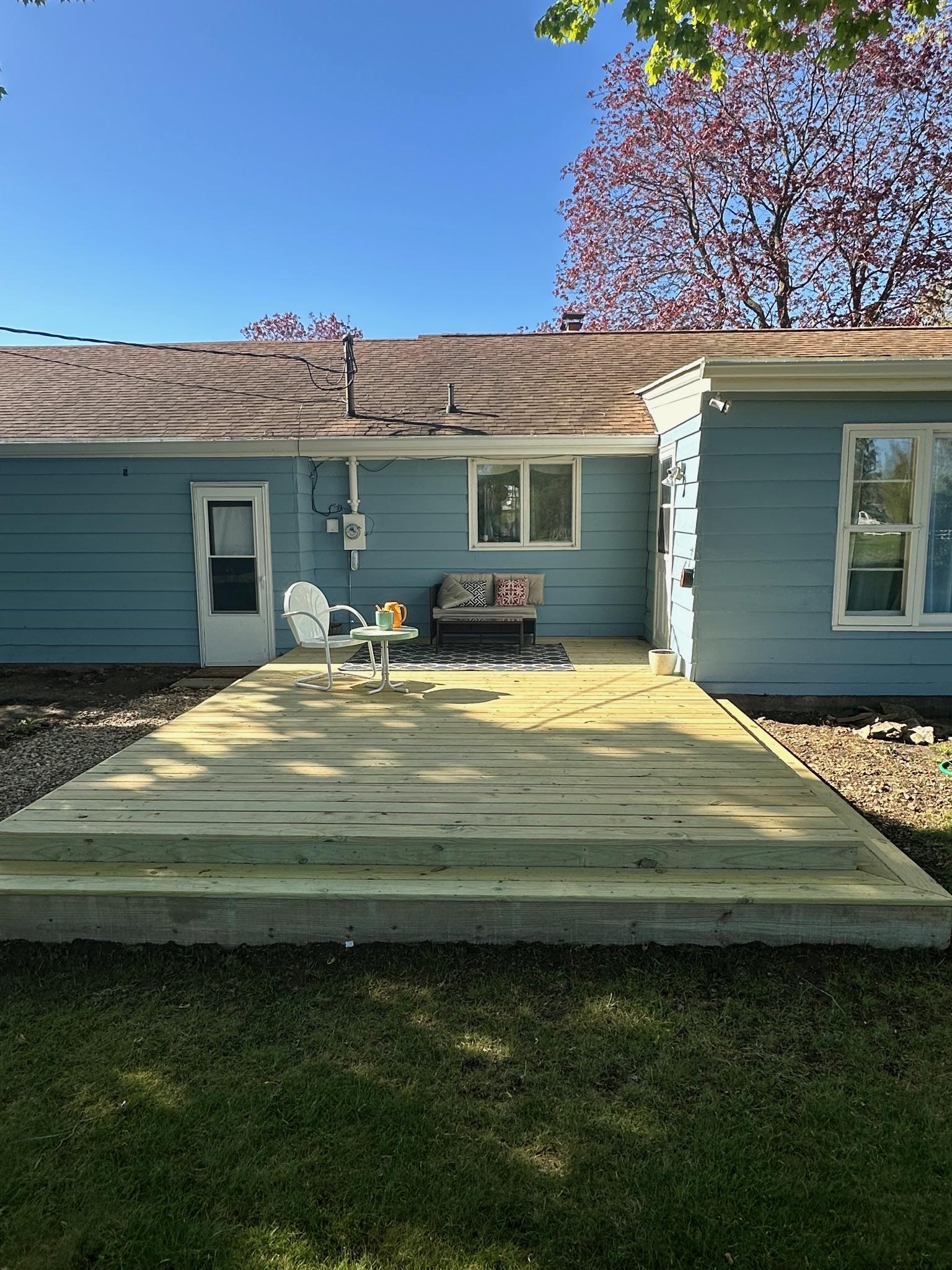 Custom ground-level pressure-treated wood patio deck with wrap-around steps built on a blue house by Precision Exterior Inc.