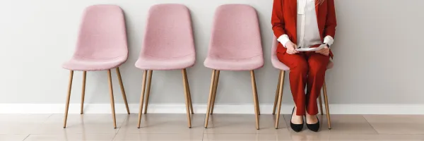 Four pink chairs with one chair occupied by a woman in a red suit waiting fir a job interview.