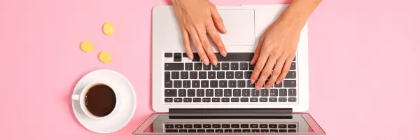 Close up of hands working on a laptop with a coffee to the side on a pink table.