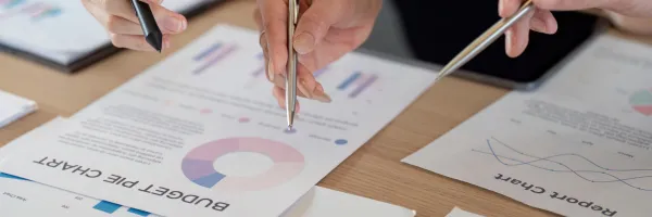 Close up of two hand, each holding a pen and pointing at business reports on sheets of paper on a table.