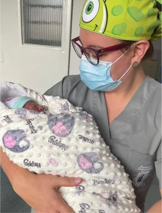 Verónica Villa con uniforme médico y mascarilla, sosteniendo a un recién nacido envuelto en una manta.