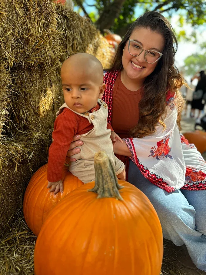Dra. Verónica Villa sonriendo junto a su bebé entre calabazas y pacas de heno en 2024.