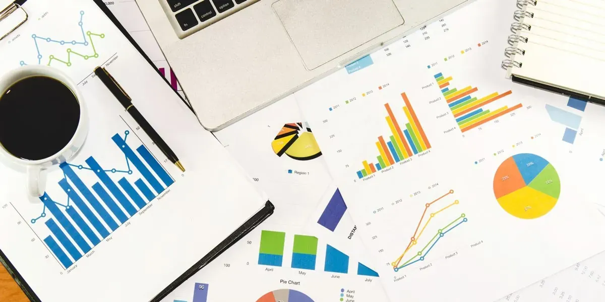 Overhead view of business reports featuring colorful bar charts, line graphs, and pie charts on a desk with a laptop, coffee cup, pen, and notebook.