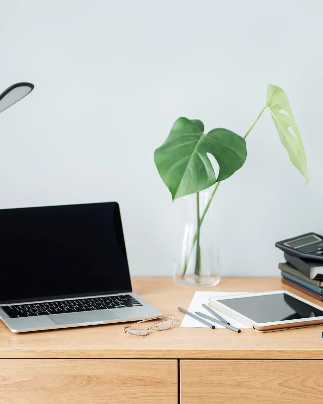 Minimalist home office desk with laptop, tablet, glasses, and monstera plant in glass vase
