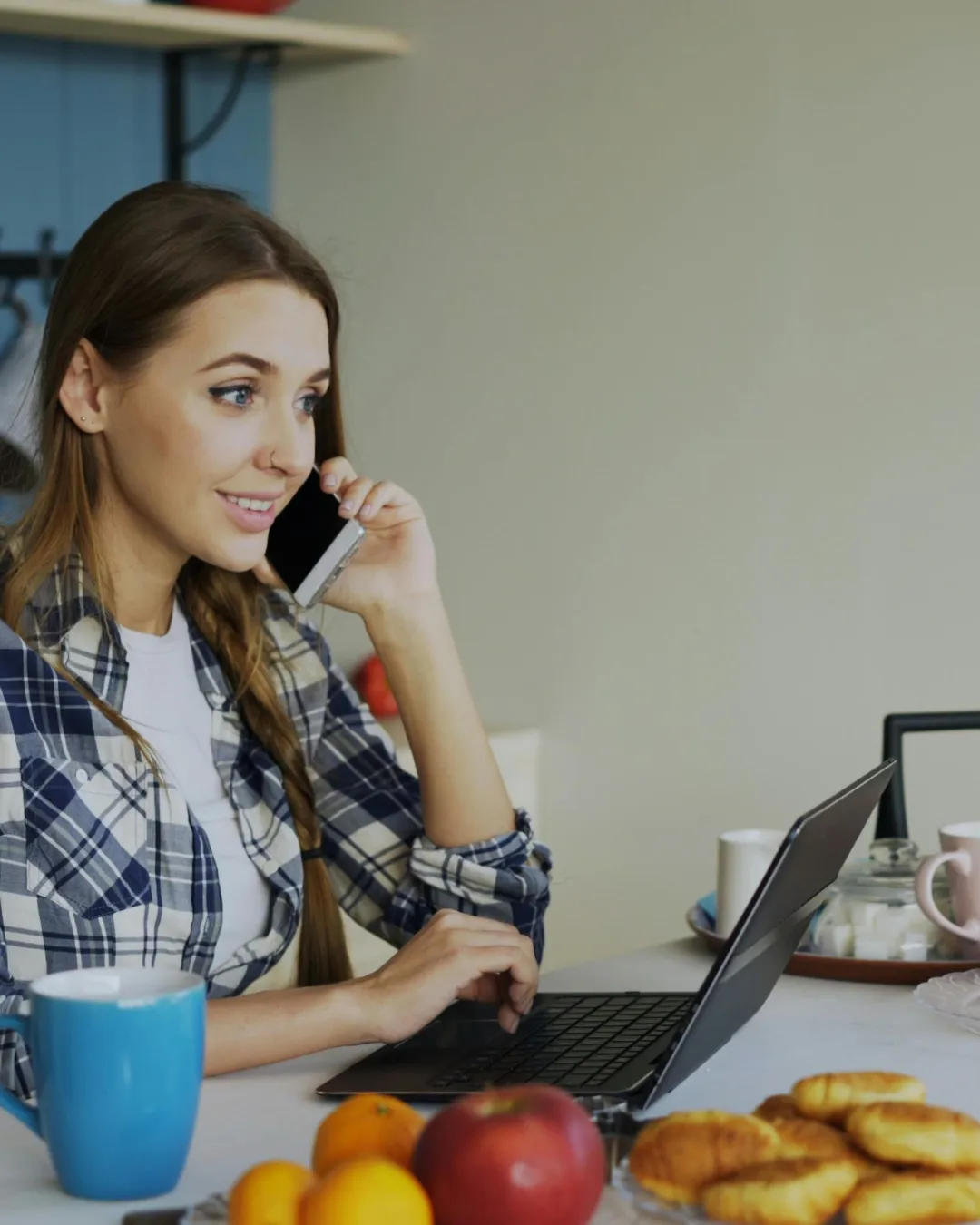 Woman working on laptop while talking on phone at kitchen table with coffee and breakfast