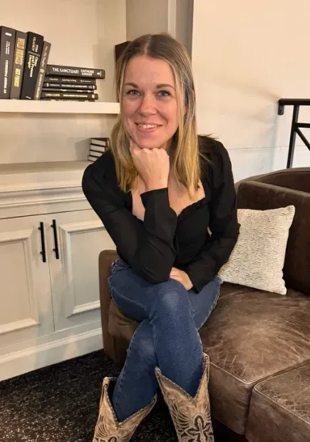 A smiling woman with shoulder-length blonde hair sits on a brown leather couch in a cozy, modern setting. She wears a black long-sleeve top, blue jeans, and intricately designed cowboy boots. Her chin rests on her hand, and she looks directly at the camera. Behind her, a white cabinet with books neatly arranged on the shelves adds a stylish touch to the background. A decorative pillow rests on the couch beside her.
