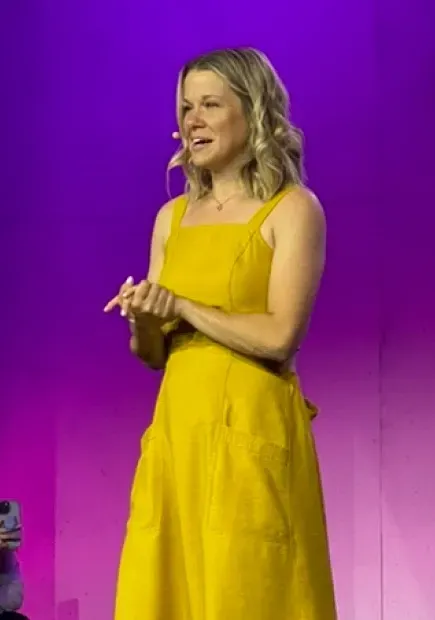A woman on stage delivering a speech, wearing a headset microphone, with a confident and engaging expression against a vibrant purple background.