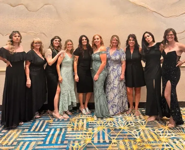**Alt Text:** A group of ten elegantly dressed women pose together in a formal setting, wearing a mix of black and pastel-colored evening gowns. They stand on a patterned carpet against a textured wall, exuding confidence and style. Some women smile warmly, while others strike playful poses, showcasing their unique personalities.