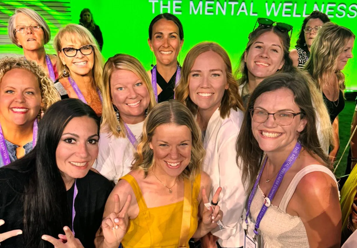 A group of smiling women pose together at a mental wellness event, wearing lanyards and standing in front of a bright green backdrop with the words "THE MENTAL WELLNESS" partially visible. The woman in the foreground, wearing a yellow dress, holds up peace signs, exuding excitement and positivity. The attendees display a mix of expressions, and the setting is a professional conference or seminar focused on wellness.