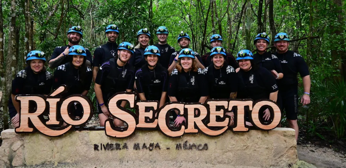A group of adventurers wearing black wetsuits and blue helmets with headlamps pose together behind a large wooden sign that reads "RÍO SECRETO" in the lush green jungle of Riviera Maya, Mexico. They are smiling and appear ready for an underground cave exploration experience. The background features dense tropical foliage, enhancing the sense of adventure.