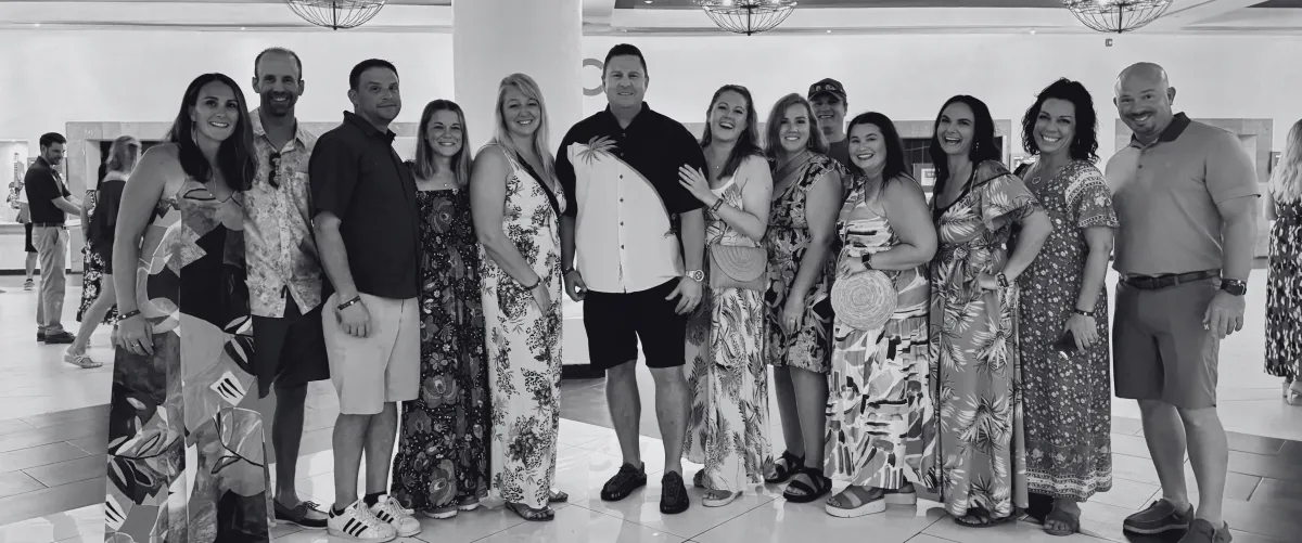 A group of twelve smiling people posing together in a well-lit indoor space. They are dressed in a mix of casual and semi-formal attire, with many wearing colorful floral-patterned dresses and shirts. The setting appears to be a hotel lobby or event venue, with modern lighting and a polished tile floor. The group stands closely together, showing camaraderie and happiness.