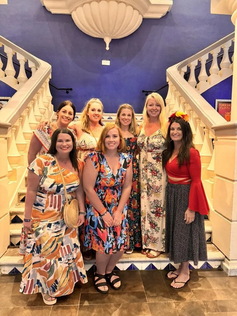 A group of seven women pose together on a grand staircase with ornate white railings and a deep blue background. They are dressed in colorful, elegant dresses with floral and patterned designs, radiating warmth and joy. One woman wears a vibrant red top with flared sleeves and a flower crown, adding a festive touch. The setting has a Mediterranean or Spanish-style aesthetic, with decorative architectural details.