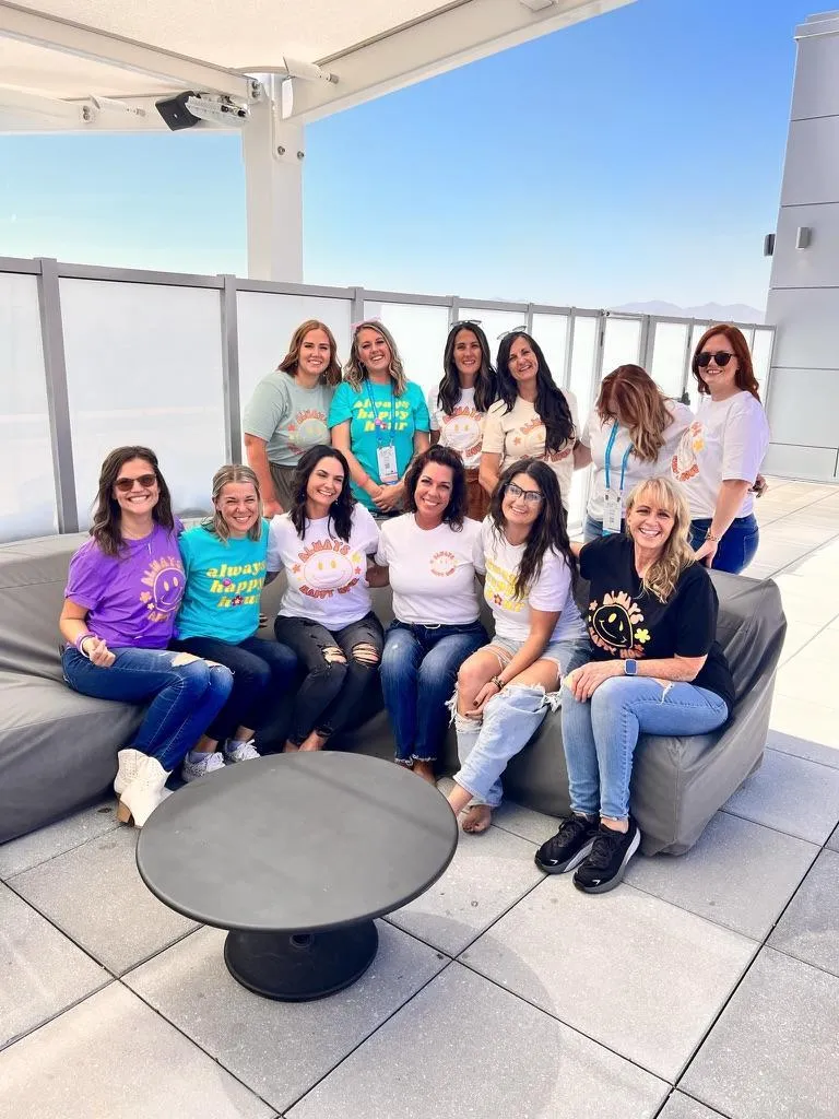 A group of twelve smiling women pose together on a rooftop lounge under a clear blue sky. They are dressed in colorful t-shirts featuring "Always Happy Hour" designs, paired with jeans and casual shoes. Some are sitting on a gray outdoor sofa while others stand behind them, radiating a sense of camaraderie and positivity. The setting is modern, with sleek furnishings and a beautiful outdoor view.