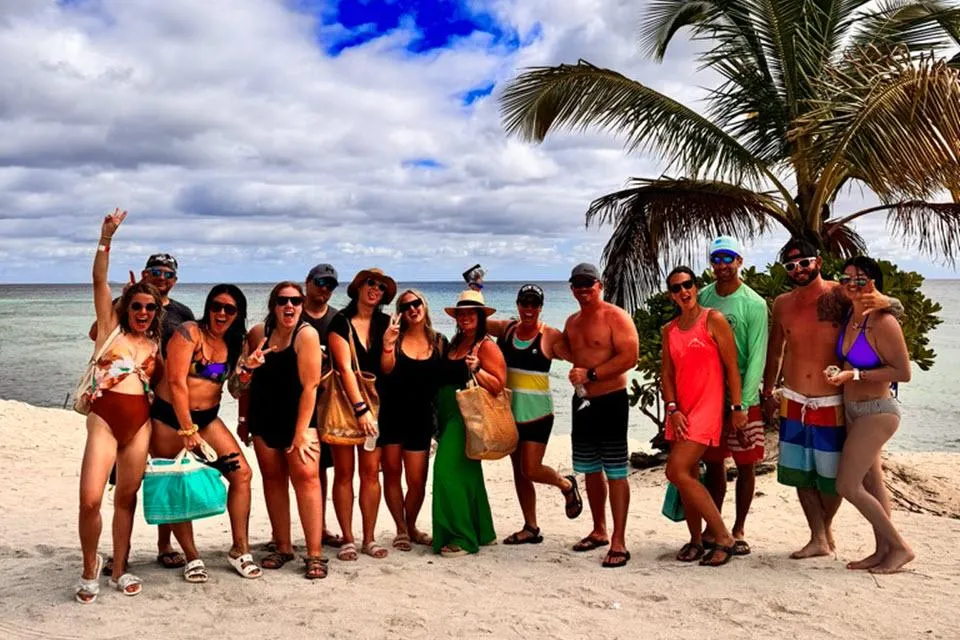**Alt Text:** A lively group of people poses together on a sandy beach with the ocean and a palm tree in the background. They are dressed in swimsuits, beachwear, and sunglasses, with some holding beach bags and drinks. Many are smiling, making peace signs, and expressing excitement, creating a fun and tropical vacation vibe. The sky is partly cloudy, adding to the picturesque beach setting.